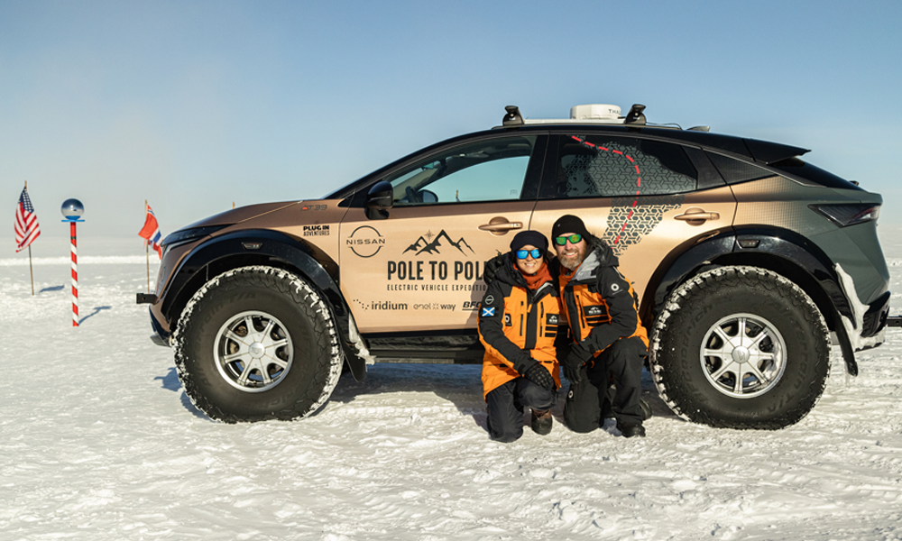 Chris and Julie kneel in front of the parked Nissan Expedition Ariya next to the pole marking the Ceremonial South Pole.